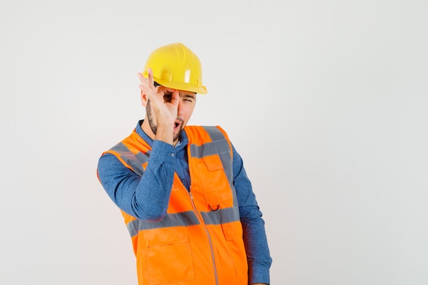 Jovem construtor mostrando sinal ok no olho na camisa, colete, capacete e olhando curioso, vista frontal.