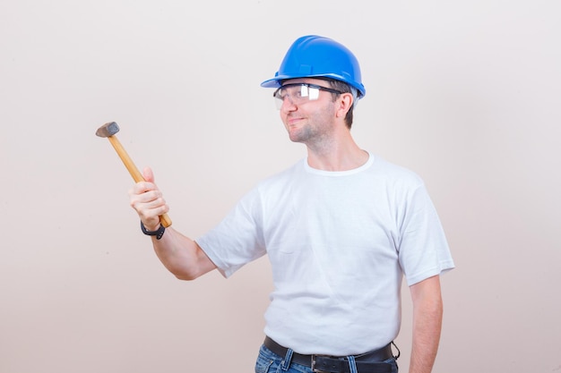 Jovem construtor olhando para o martelo de camiseta, jeans, capacete e parecendo esperançoso