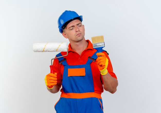 Foto grátis jovem construtor pensando vestindo uniforme e capacete de segurança segurando o rolo de pintura e olhando para o pincel na mão