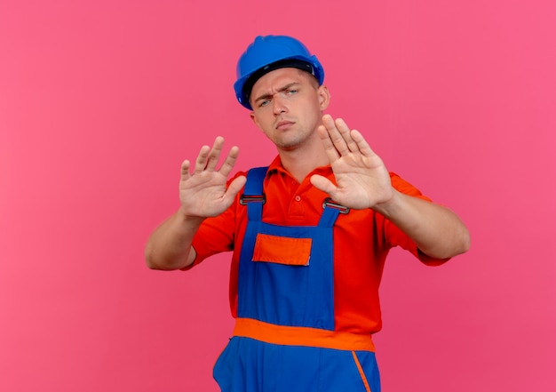 Jovem construtor rigoroso usando uniforme e capacete de segurança mostrando gesto de parada em rosa