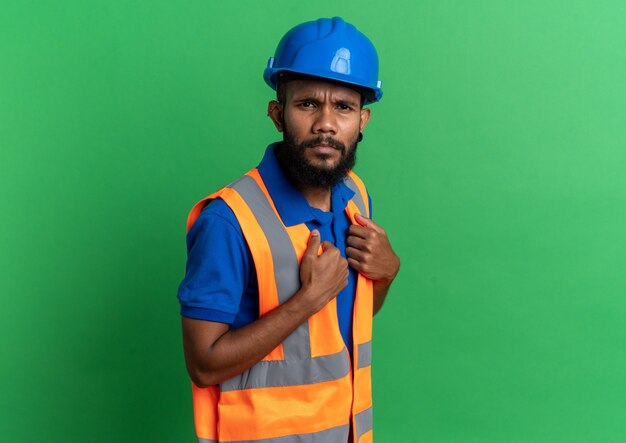 jovem construtor sem noção, de uniforme, com capacete de segurança, olhando para a frente, isolado em uma parede verde com espaço de cópia