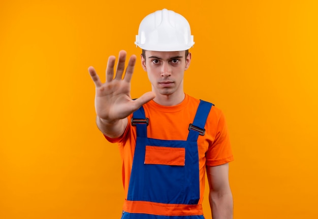 Jovem construtor vestindo uniforme de construção e capacete de segurança, fazendo stop com a mão esquerda