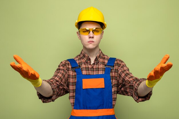 Jovem construtor vestindo uniforme e luvas com óculos isolados na parede verde oliva