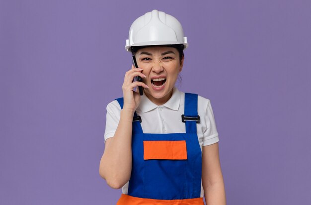 Jovem construtora asiática irritada com capacete de segurança branco gritando com alguém no telefone