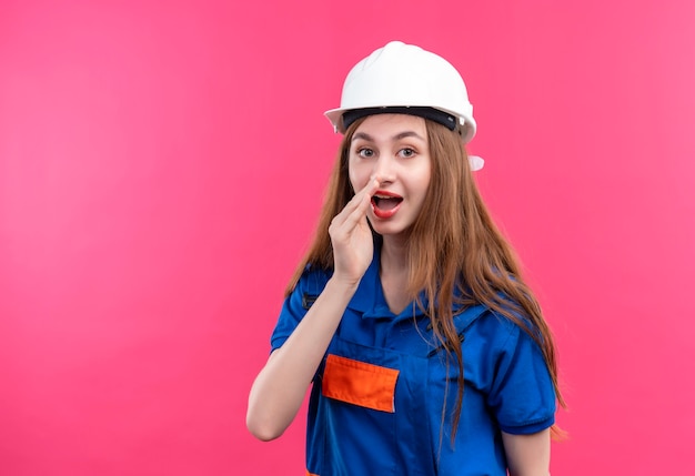 Jovem construtora com uniforme de construção e capacete de segurança, contando um segredo com a mão perto da boca, em pé sobre a parede rosa