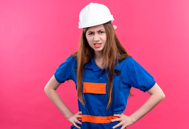 Jovem construtora com uniforme de construção e capacete de segurança olhando com cara de raiva em pé sobre a parede rosa