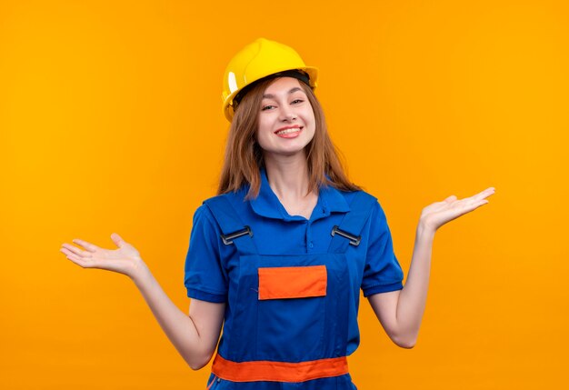 Jovem construtora com uniforme de construção e capacete de segurança olhando estendendo os braços para os lados sorrindo em pé