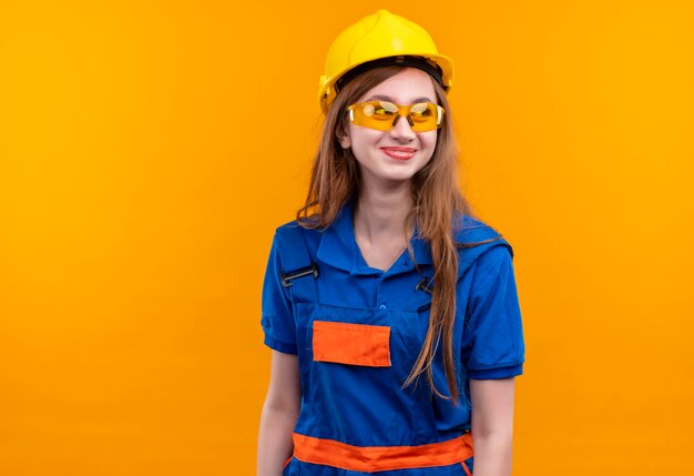 Jovem construtora com uniforme de construção e capacete de segurança olhando para o lado e sorrindo alegremente em pé