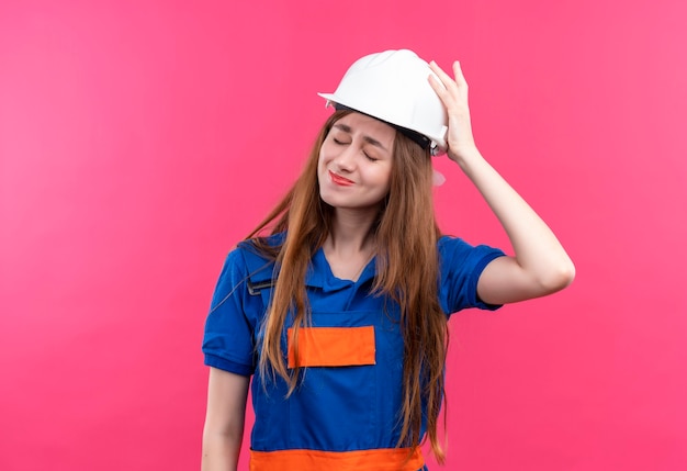 Jovem construtora com uniforme de construção e capacete de segurança, parecendo confusa com a mão na cabeça por engano em pé sobre a parede rosa