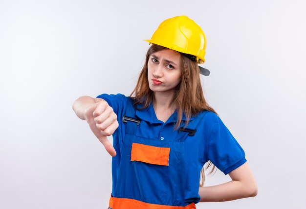 Jovem construtora com uniforme de construção e capacete de segurança, parecendo descontente mostrando os polegares para baixo em pé sobre uma parede branca