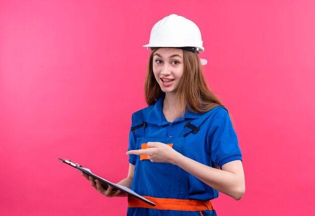 Jovem construtora com uniforme de construção e capacete de segurança segurando uma prancheta apontando com o dedo para ela sorrindo em pé sobre a parede rosa