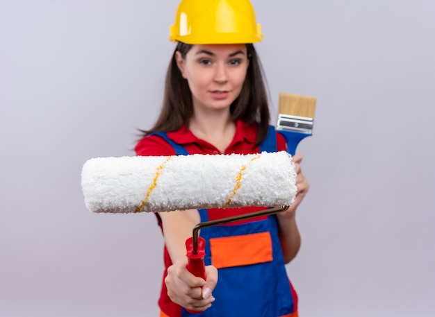 Foto grátis jovem construtora confiante segurando rolo de pintura e pincel no fundo branco isolado