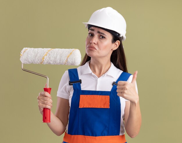 Foto grátis jovem construtora de uniforme, segurando a escova giratória e aparecendo o polegar isolado na parede verde oliva, pensando