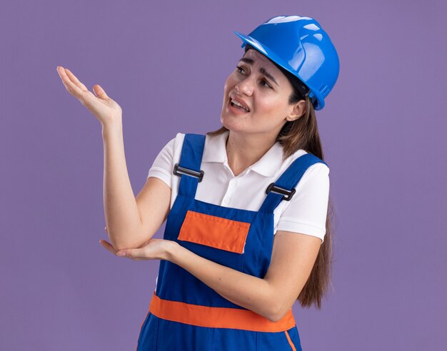 Jovem construtora descontente olhando para o lado, em pontos uniformes, com a mão na lateral isolada na parede roxa