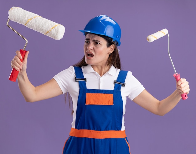 Jovem construtora insatisfeita de uniforme segurando um mini rolo de tinta e olhando para a escova de rolo na mão, isolada na parede roxa