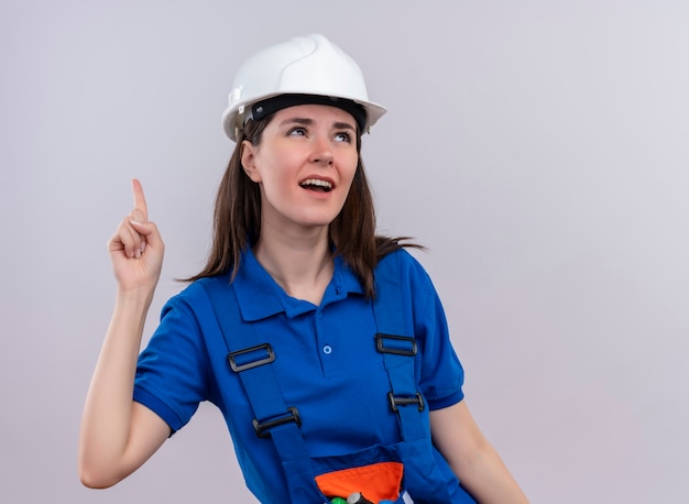 Jovem construtora irritada com capacete de segurança branco e uniforme azul aponta para cima e olha para um fundo branco isolado com espaço de cópia