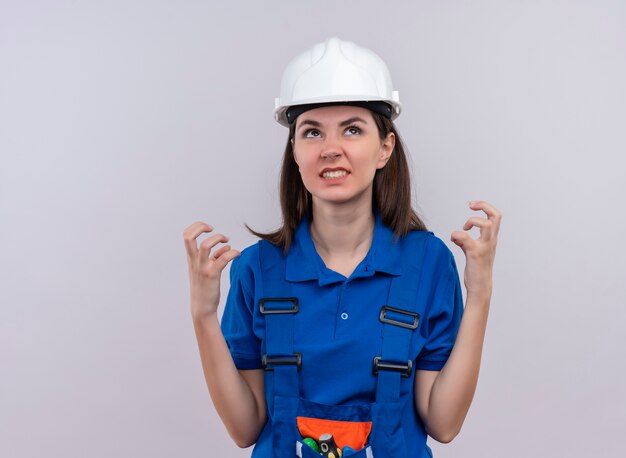 Jovem construtora irritada com capacete de segurança branco e uniforme azul levanta as mãos e olha para cima, em um fundo branco isolado com espaço de cópia