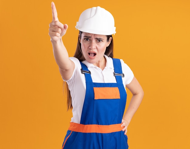 Foto grátis jovem construtora rígida em pontos uniformes para a câmera, colocando a mão no quadril isolado na parede laranja