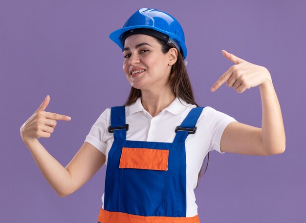 Jovem construtora sorridente em uniforme aponta para si mesma, isolada na parede roxa