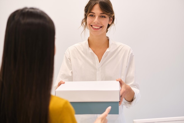 Jovem consultor de moda sorridente alegremente dando sapatos na caixa para o cliente na loja moderna