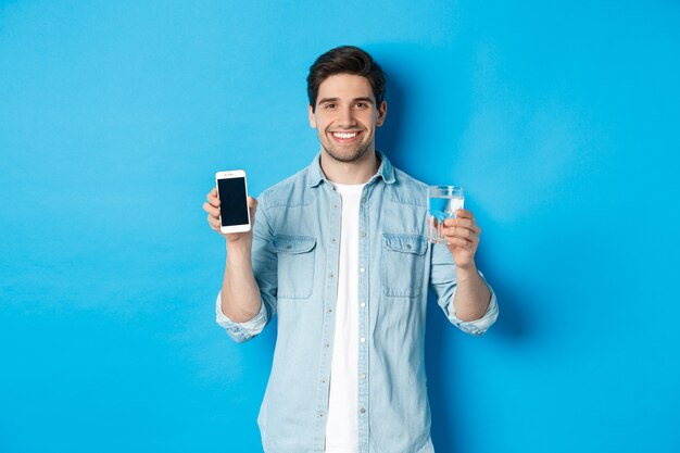 Jovem controla o equilíbrio da água com o aplicativo do smartphone, mostrando o aplicativo da tela do celular e sorrindo, de pé sobre o fundo azul