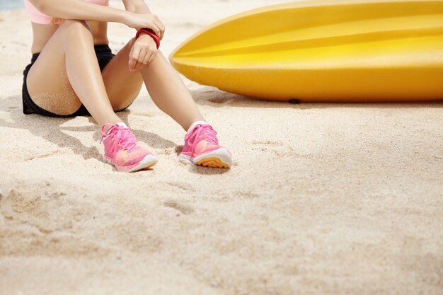 Jovem corredor feminino com bela pele bronzeada no sportswear e tênis, sentado na areia perto do barco amarelo e relaxante após treinamento físico intensivo ao ar livre, preparando-se para a maratona