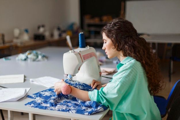 Jovem costureira com cabelo encaracolado escuro em camisa colorida pensativamente usando máquina de costura na oficina de costura moderna