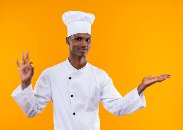 Jovem cozinheira afro-americana com uniforme de chef gesticula bem e mantém a mão reta isolada na parede laranja
