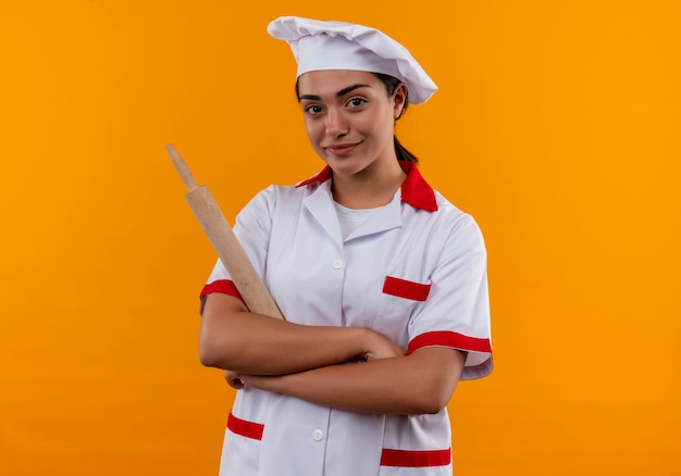 Jovem cozinheira caucasiana confiante com uniforme de chef cruza os braços e segura o rolo de massa isolado em um fundo laranja com espaço de cópia