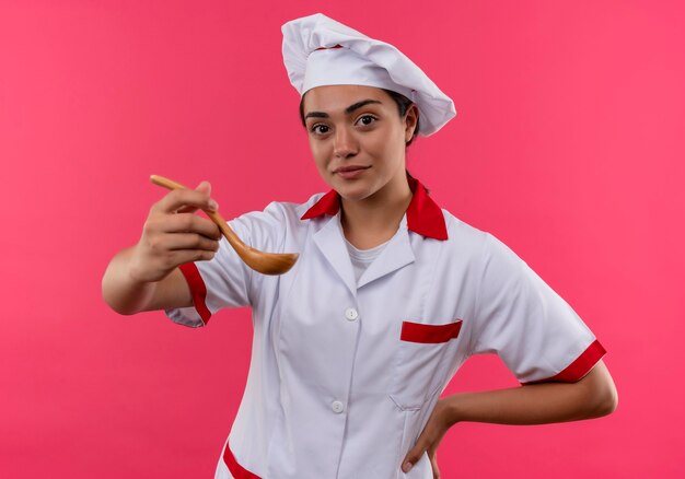 Foto grátis jovem cozinheira caucasiana confiante com uniforme de chef segura uma colher de pau e coloca a mão na cintura, isolada na parede rosa com espaço de cópia