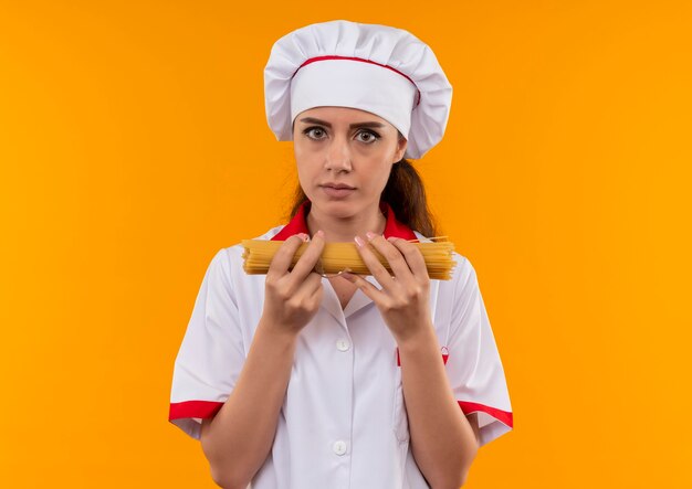 Jovem cozinheira caucasiana confiante com uniforme de chef segurando um monte de espaguete com as duas mãos isolado na parede laranja com espaço de cópia