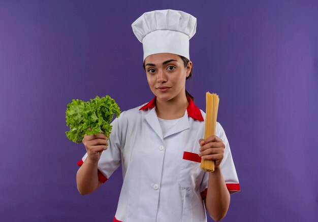 Jovem cozinheira caucasiana confiante em uniforme de chef com salada e molho de espaguete isolado na parede violeta com espaço de cópia
