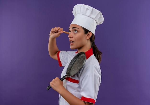 Foto grátis jovem cozinheira caucasiana irritada com uniforme de chef fica de lado e segura a frigideira e a colher de pau, isoladas no fundo violeta com espaço de cópia