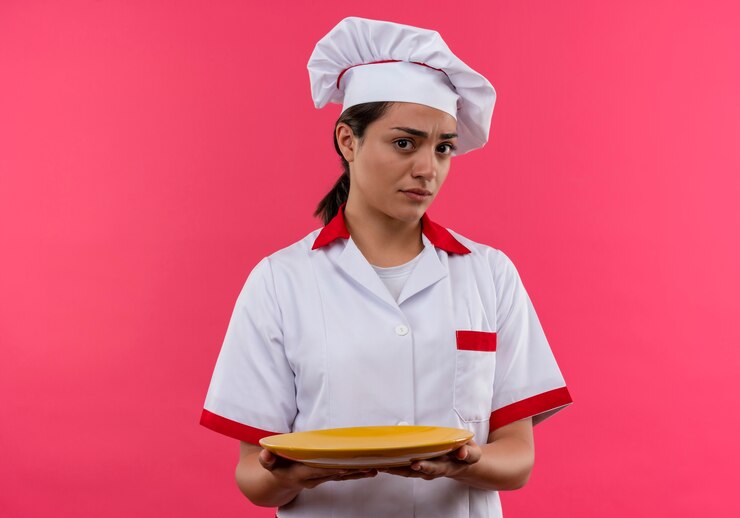 Foto grátis jovem cozinheira caucasiana irritada com uniforme de chef segura o prato com as duas mãos isoladas no fundo rosa com espaço de cópia