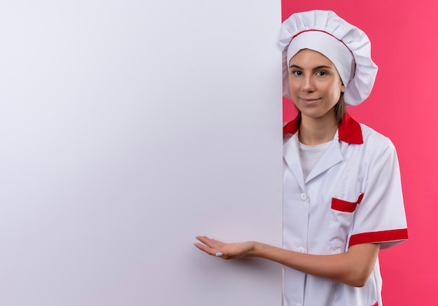 Jovem cozinheira caucasiana satisfeita com uniforme de chef fica atrás e aponta para uma parede branca em rosa com espaço de cópia