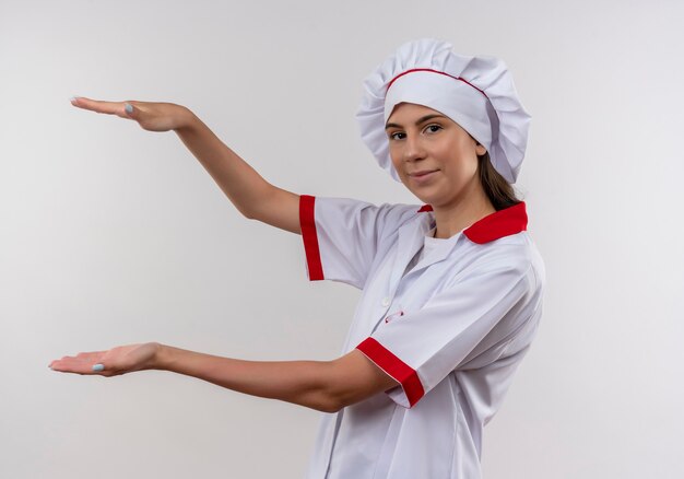 Jovem cozinheira caucasiana satisfeita com uniforme de chef fica de lado e finge segurar algo em branco com espaço de cópia