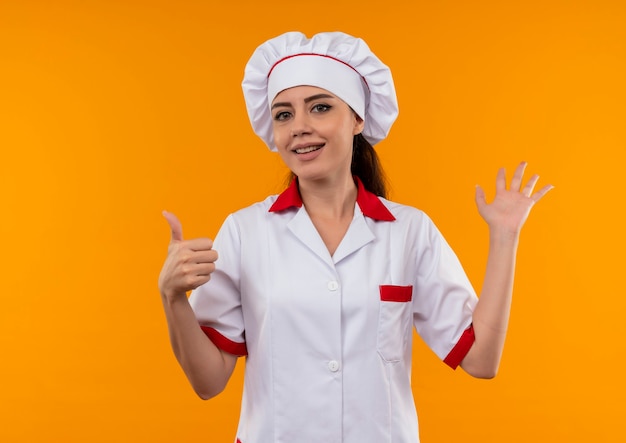 Jovem cozinheira caucasiana satisfeita com uniforme de chef levanta a mão e levanta a mão isolada na parede laranja com espaço de cópia