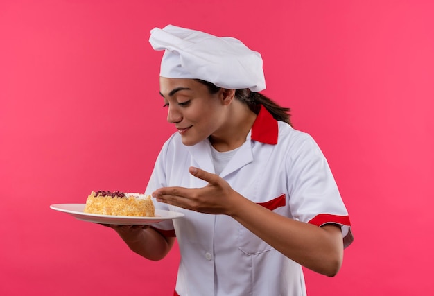 Foto grátis jovem cozinheira caucasiana satisfeita com uniforme de chef segura e olha para o bolo no prato isolado na parede rosa com espaço de cópia