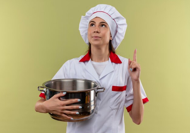 Jovem cozinheira caucasiana satisfeita com uniforme de chef segurando a panela e aponta para cima