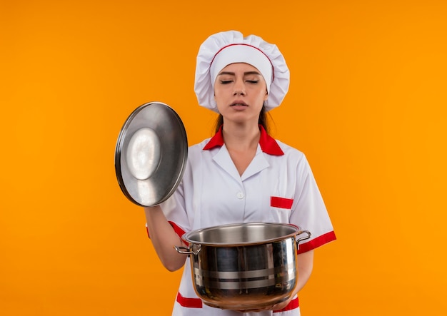 Jovem cozinheira caucasiana satisfeita com uniforme de chef segurando a panela e fingindo cheirar isolado na parede laranja com espaço de cópia