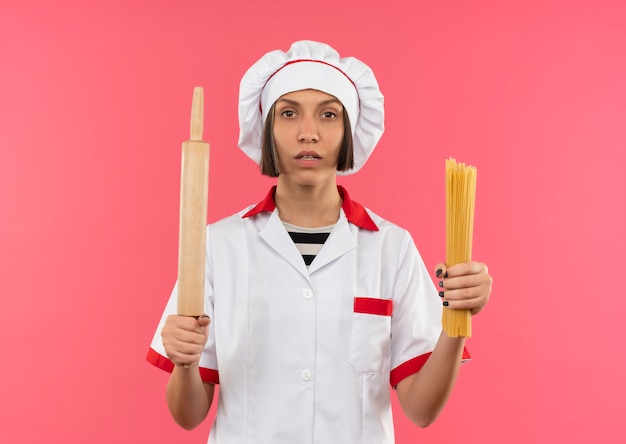 Jovem cozinheira confiante em uniforme de chef segurando macarrão espaguete e rolo de massa e parecendo isolada em rosa