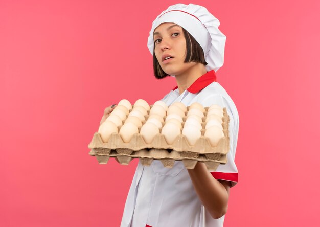 Jovem cozinheira confiante em uniforme de chef segurando uma caixa de ovos isolada em rosa com espaço de cópia