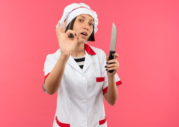 Jovem cozinheira confiante em uniforme de chef, segurando uma faca e fazendo sinal de ok, isolada em rosa com espaço de cópia