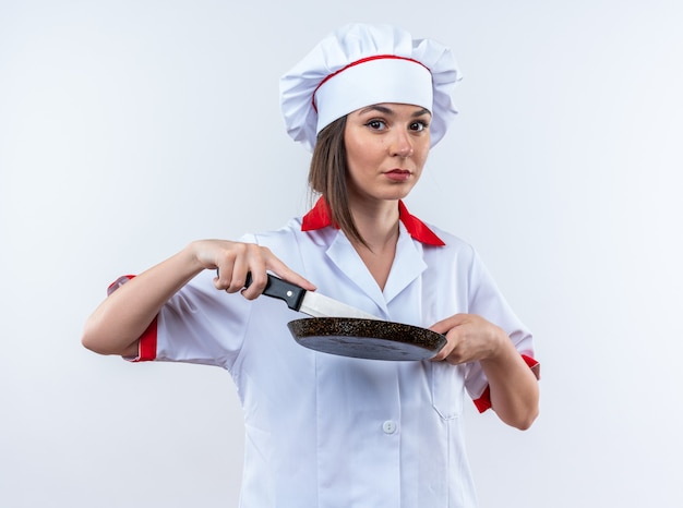 Jovem cozinheira confiante vestindo uniforme de chef segurando uma frigideira com uma faca isolada no fundo branco