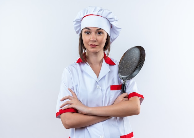 Foto grátis jovem cozinheira confiante vestindo uniforme de chef segurando uma frigideira cruzando as mãos isoladas na parede branca