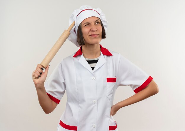 Jovem cozinheira confusa em uniforme de chef, segurando o rolo de massa e colocando a mão na cintura, olhando para cima isolado no branco