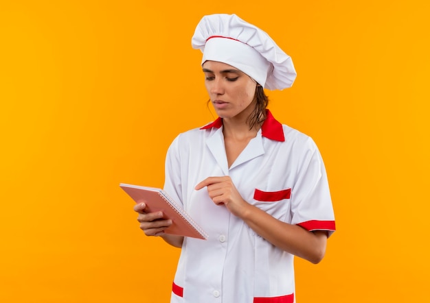 Jovem cozinheira confusa vestindo uniforme de chef segurando e olhando para o caderno na parede amarela isolada com espaço de cópia