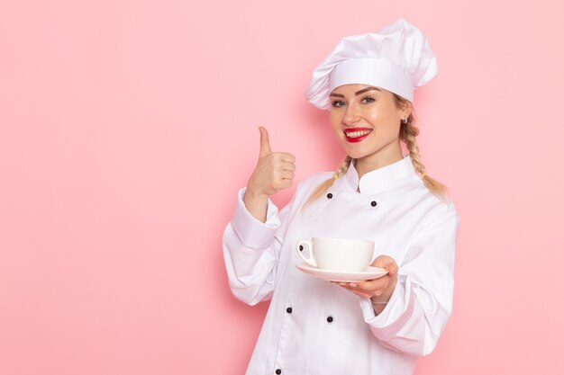 Jovem cozinheira de terno branco segurando uma xícara de café com um leve sorriso na cozinha do espaço rosa