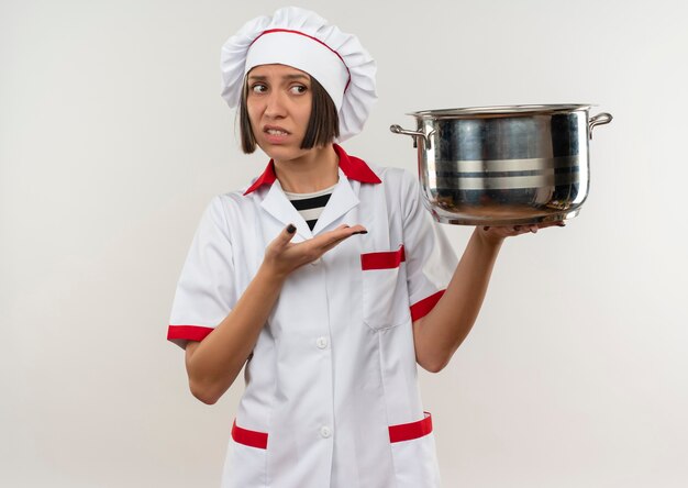 Jovem cozinheira descontente em uniforme de chef segurando e apontando com a mão para a panela, olhando para o lado isolado no fundo branco com espaço de cópia