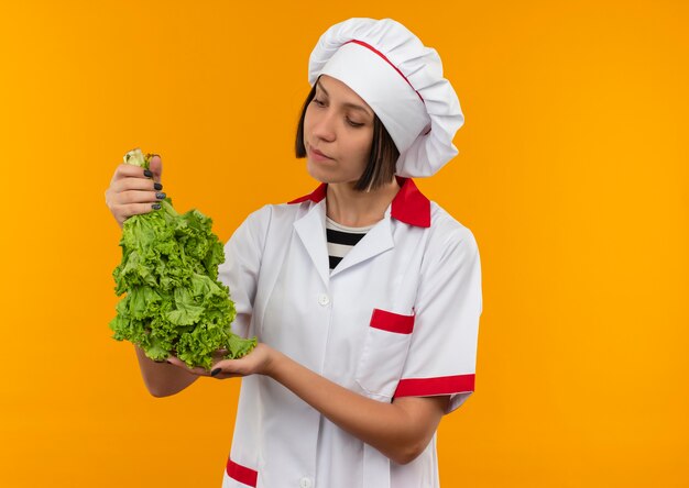 Jovem cozinheira em uniforme de chef segurando e olhando para alface isolada em um fundo laranja com espaço de cópia